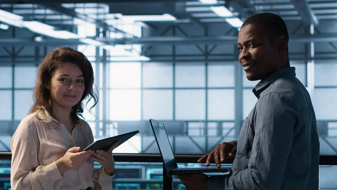 Улыбающиеся администраторы используют ноутбук в дата-центре, управляя виртуализацией серверов.